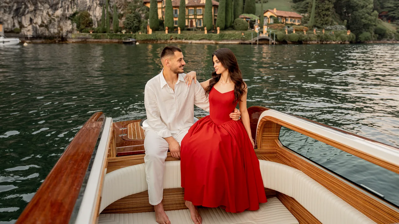 A couple sitting on the open deck of a wooden boat on Lake Como, with a lakeside villa behind them.